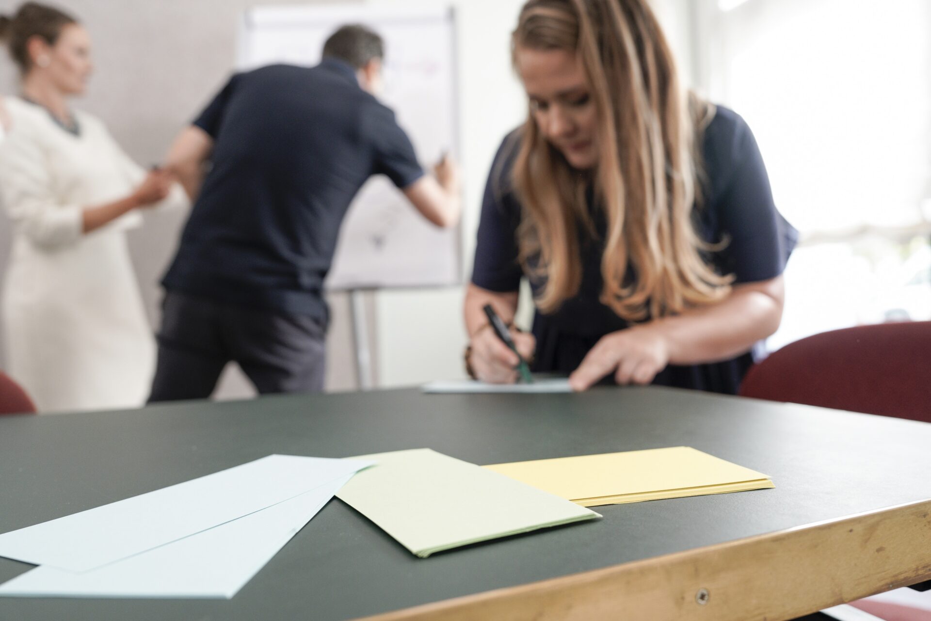 Frau schreibt am Tisch, Team arbeitet im Hintergrund